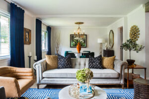A stylish living room blending Mid-Century Modern and Maximalist design, featuring a tufted gray sofa with patterned pillows, a leather armchair, bold blue rug, and eclectic decorative pieces.