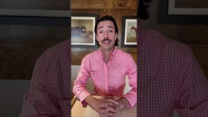 JohnRoss seated in a wood-paneled room wearing a pink checkered shirt, sharing emotional interior design tips on how your home reflects your personal life story.