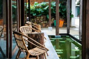 Rattan chairs arranged around wooden stump tables beside a reflective water feature with lily pads in a natural, minimalist patio setting.