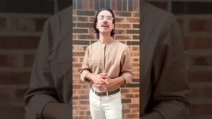 John Ross stands against a warm-toned brick wall, wearing a taupe shirt and glasses, sharing insights on the value and longevity of antique furniture in interior design.